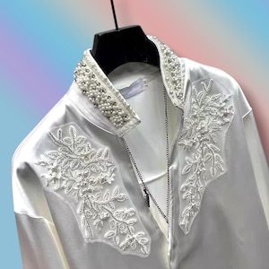 Op de afbeelding: Witte satijnen blouse met een kraag van kralen en bloemenborduurwerk op de voorkant. De blouse heeft een lange zilveren ketting en hangt aan een zwarte hanger. Het borduurwerk en de kraag zijn versierd met parels.