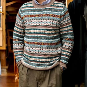 May include: A patterned jumper in shades of teal, brown, and cream. The jumper features a crew neck and long sleeves. The design includes geometric and striped patterns. A light blue collared shirt is worn under the jumper, paired with brown trousers.