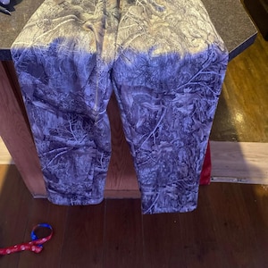May include: Camouflage-patterned sweatpants with an elastic waistband. The pants feature a forest-like design in shades of brown, gray, and white. The fabric appears to be a soft, comfortable material, suitable for casual wear.