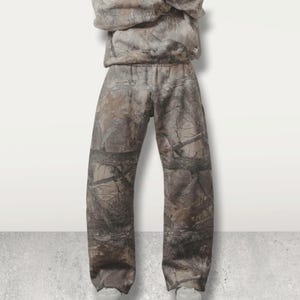 May include: A two-piece camouflage outfit, featuring a hooded sweatshirt and matching pants. The pants have an elastic waistband. The camouflage pattern incorporates shades of brown, tan, and green, mimicking a forest environment. The outfit is made of a soft material.