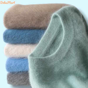 Pode incluir: Uma pilha de camisolas macias e fofas em várias cores, incluindo bege, azul e cinza. Uma camisola verde clara é parcialmente visível no lado direito. As camisolas parecem ser feitas de um material felpudo.