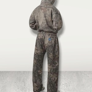 May include: A two-piece camouflage outfit, including a hooded sweatshirt and matching pants. The outfit features a brown, green, and gray woodland pattern. The pants have a back pocket with a blue square patch.