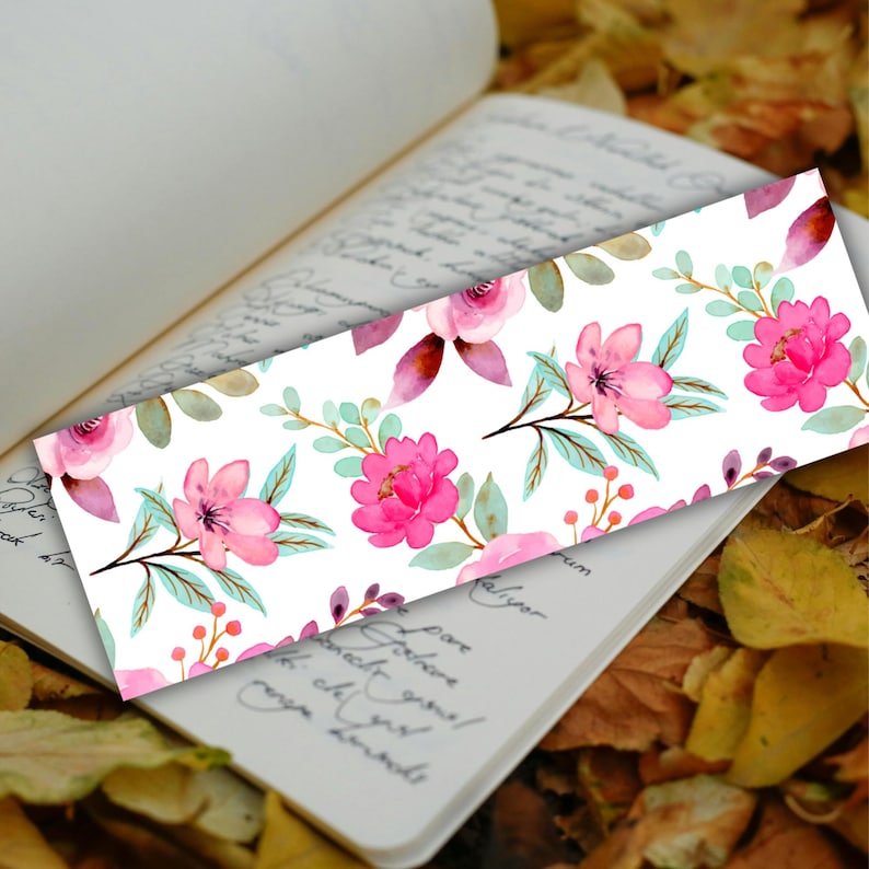 May include: A floral bookmark featuring a watercolor design of pink flowers, green leaves, and purple accents on a white background. The bookmark is rectangular and rests on an open book with handwritten text, surrounded by autumn leaves.