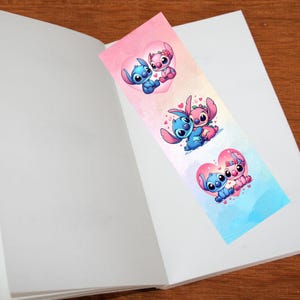May include: A colorful bookmark featuring cartoon characters in heart-shaped designs. The bookmark has a gradient background of pink and blue. The characters are blue and pink, with large eyes and expressive faces. The bookmark is placed inside an open book.