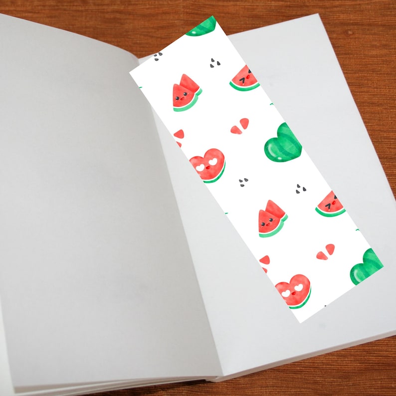 May include: A white bookmark with a repeating pattern of watermelon slices, heart-shaped watermelons, and green heart shapes. The bookmark is placed inside an open book with white pages, resting on a wooden surface.