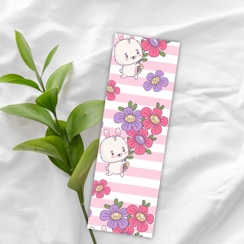 May include: A bookmark featuring a cartoon bunny holding flowers, set against a pink and white striped background. The design includes purple and pink flowers. The bookmark is a whimsical accessory for book lovers.