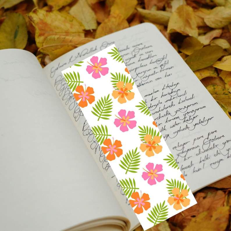 May include: A white bookmark with a repeating pattern of pink and orange hibiscus flowers and green leaves. The bookmark is placed in an open book with handwritten text on the pages. The background features autumn leaves.