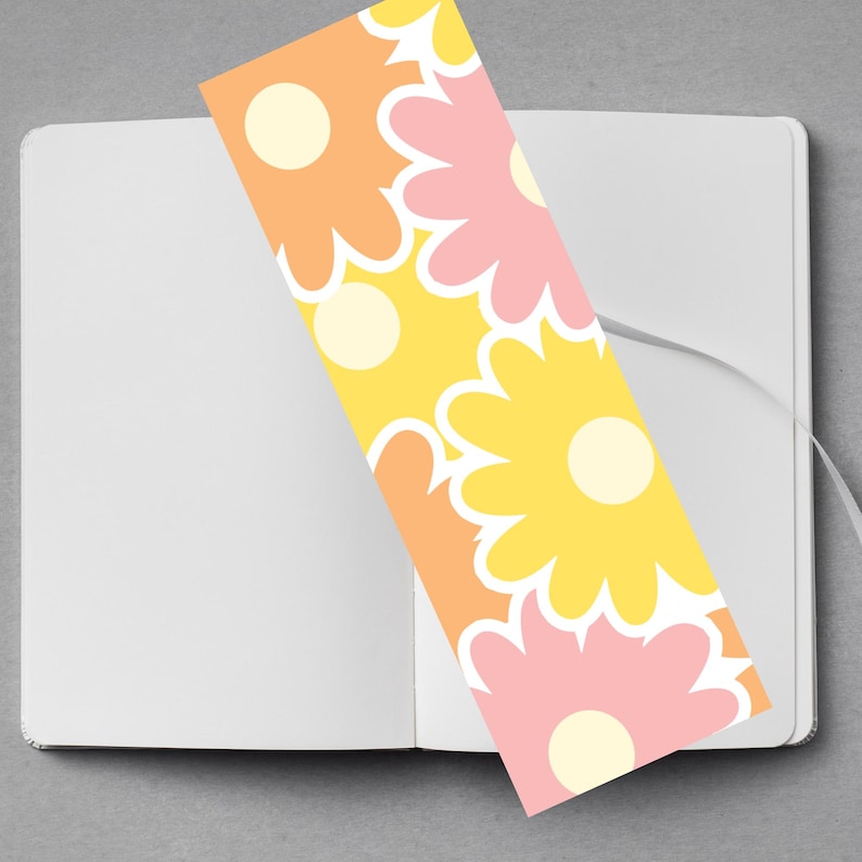 May include: A floral bookmark with a retro design rests inside an open notebook. The bookmark features large, stylized flowers in shades of yellow, orange, and pink with white accents. The notebook is white with a gray background.