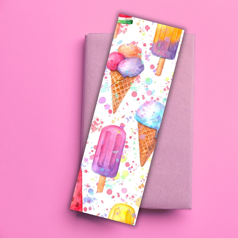 May include: A watercolor bookmark featuring colorful ice cream cones and popsicles on a white background with paint splatters. The bookmark is resting on a closed book, with a pink background.