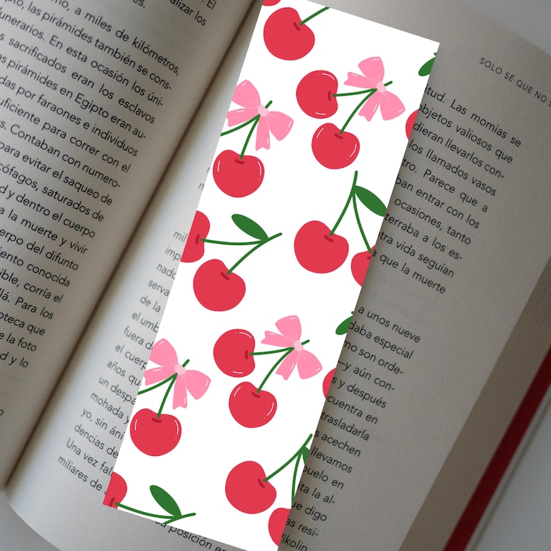 May include: A white bookmark with a pattern of red cherries, green stems, and pink bows. The bookmark is placed in an open book, with text visible on the pages. The design is playful and suitable for book lovers.