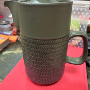 May include: A stoneware coffee press in dark olive green. The lower section has a textured, woven design. The lid and handle are a darker brown. The press is on a red surface. The height is approximately 18 cm.