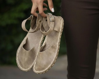 Puede incluir: Un par de sandalias de cuero color topo con correas en el tobillo y cierres de hebilla. Las sandalias tienen un diseño cosido alrededor de los bordes. El calzado está diseñado con la punta abierta y tacón bajo.