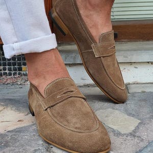 May include: Brown suede loafers with a penny strap detail. The shoes have a rounded toe and a stitched design around the edges. The soles are a light brown color.
