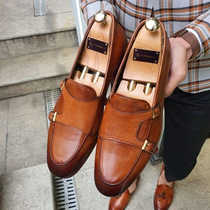 Könnte beinhalten: Ein Paar braune Leder-Monk-Strap-Schuhe mit goldfarbenen Schnallen. Die Schuhe werden von einer Person gehalten, die das polierte Leder und die hölzernen Schuhspanner im Inneren zeigt. Die Schuhe haben ein klassisches Design.