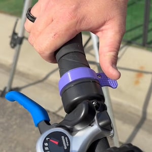 Może przedstawiać: Zbliżenie na kierownicę roweru z fioletowym dzwonkiem i niebieską dźwignią. Dzwonek ma teksturowaną dźwignię, a dźwignia ma logo Shimano i numer "1.95".