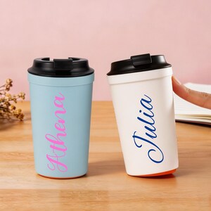 Pode incluir: Duas canecas de viagem reutilizáveis com tampas pretas. Uma caneca é azul-clara com o nome Athena escrito em letra cursiva rosa. A outra caneca é branca com o nome Julia escrito em letra cursiva azul. Ambas têm um acabamento liso.