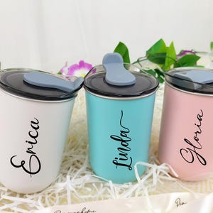 May include: Three personalized tumblers in white, teal, and pink, each with a black lid and a gray flip-top. The names "Erica", "Linda", and "Gloria" are written in cursive on the front of each tumbler. The tumblers are displayed on a bed of white paper.