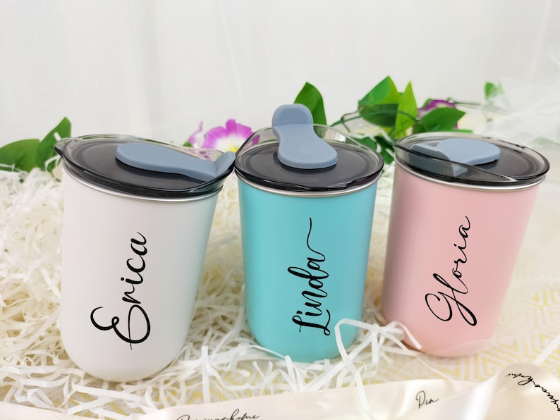 May include: Three personalized tumblers in white, teal, and pink, each with a black lid and a gray flip-top. The names "Erica", "Linda", and "Gloria" are written in cursive on the front of each tumbler. The tumblers are displayed on a bed of white paper.