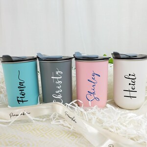 May include: Four personalized tumblers in teal, gray, pink, and white, each with a name in a contrasting color. The tumblers have black lids and are set on a bed of white paper. A white ribbon with text is in the foreground.