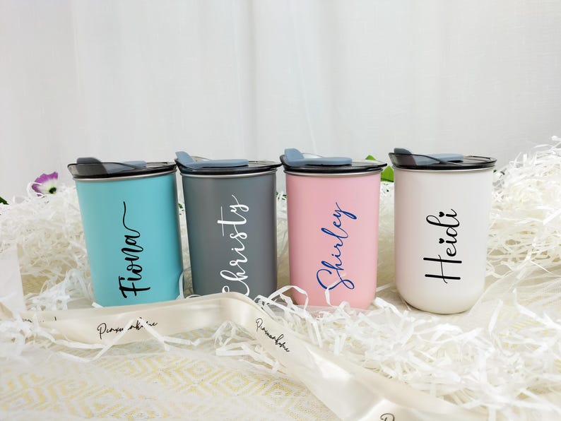 May include: Four personalized tumblers in teal, gray, pink, and white, each with a name in a contrasting color. The tumblers have black lids and are set on a bed of white paper. A white ribbon with text is in the foreground.