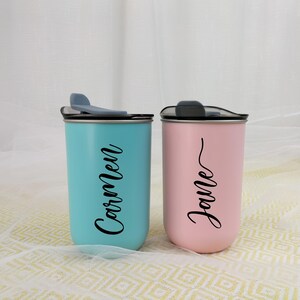 May include: Two personalized tumblers, one teal and one pink, each with a black lid and a name in a cursive font. The teal tumbler says "Carmen", and the pink tumbler says "Jane".
