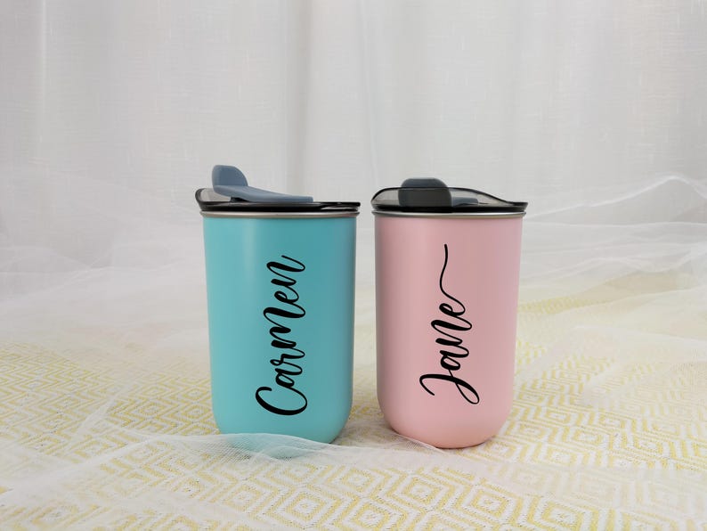 May include: Two personalized tumblers, one teal and one pink, each with a black lid and a name in a cursive font. The teal tumbler says "Carmen", and the pink tumbler says "Jane".