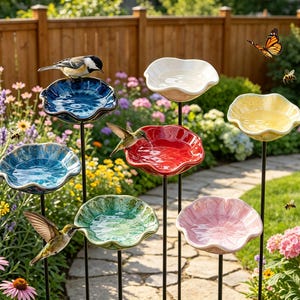 May include: A collection of six ceramic bird baths on thin black metal stakes, each featuring a scalloped flower-shaped basin in various glossy colors including blue, red, white, yellow, green, and pink, with birds and a butterfly nearby.