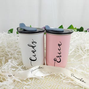 May include: Two personalized tumblers, one white and one pink, each with a name in elegant script. The tumblers have black lids with a blue slider and are set on a bed of white paper. A white ribbon with text is in the foreground.
