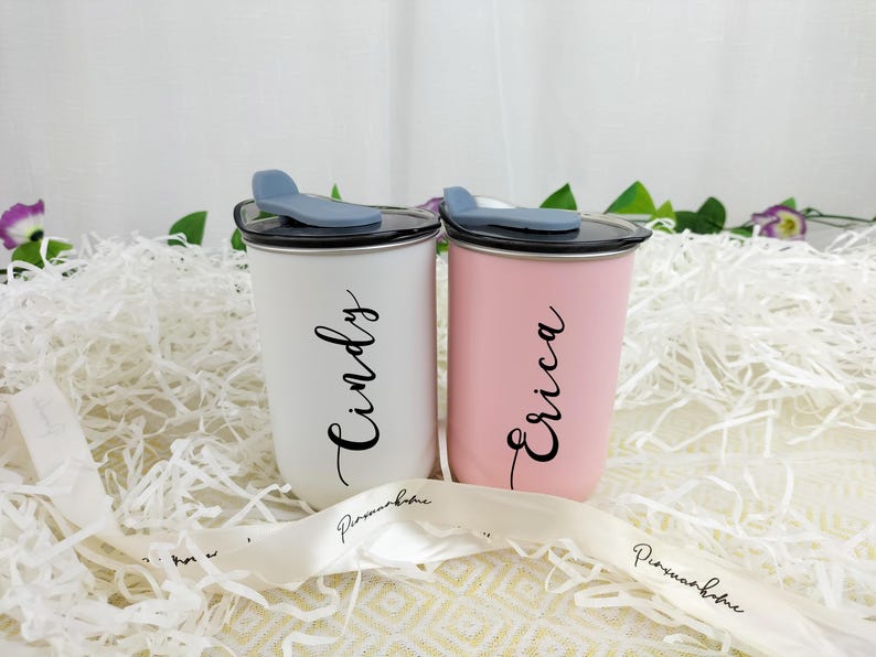 May include: Two personalized tumblers, one white and one pink, each with a name in elegant script. The tumblers have black lids with a blue slider and are set on a bed of white paper. A white ribbon with text is in the foreground.