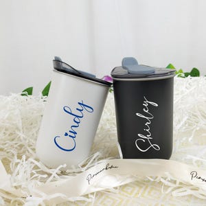 May include: Two personalized tumblers, one white and one black, with lids. The white tumbler has the name "Cindy" in blue script, and the black tumbler has the name "Shirley" in white script. They are set on white paper filler.