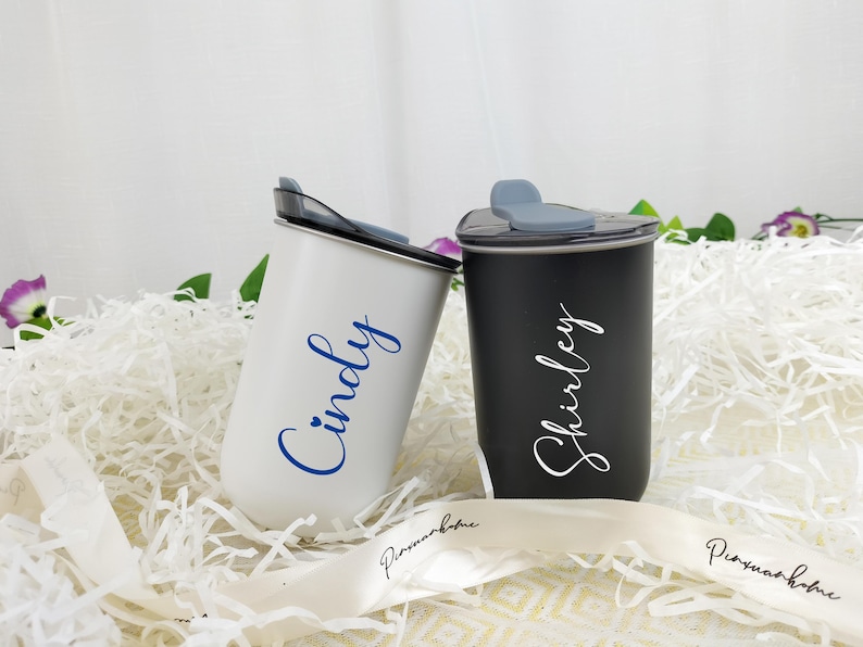 May include: Two personalized tumblers, one white and one black, with lids. The white tumbler has the name "Cindy" in blue script, and the black tumbler has the name "Shirley" in white script. They are set on white paper filler.