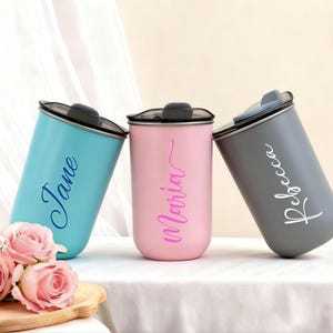 May include: Three personalized travel mugs in teal, pink, and gray, each with a name in cursive script: "Jane", "Maria", and "Rebecca". The mugs have black lids and are displayed with pink roses on a wooden tray.