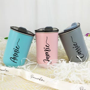 May include: Three personalized tumblers in light blue, pink, and gray. Each tumbler has a black lid and a name in a cursive font: "Annie", "Jamie", and "Carrie". The tumblers are cylindrical and sit on a bed of white paper shreds.