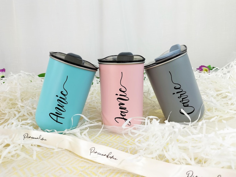 May include: Three personalized tumblers in light blue, pink, and gray. Each tumbler has a black lid and a name in a cursive font: "Annie", "Jamie", and "Carrie". The tumblers are cylindrical and sit on a bed of white paper shreds.