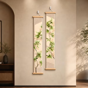 May include: Two vertical wall hanging scrolls featuring a botanical illustration of green leafy branches against a light beige textured background. Each scroll is mounted on light wood bars at the top and bottom with a white string for hanging.