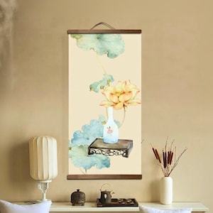 Könnte beinhalten: Ein vertikaler Wandbehang mit einem Aquarellgemälde einer Lotusblume und Blättern in Gelb-, Blau- und Grüntönen. Eine kleine, weiße Vase mit floralem Design steht auf einem dunklen Holzregal. Das Kunstwerk ist mit Holzleisten gerahmt.