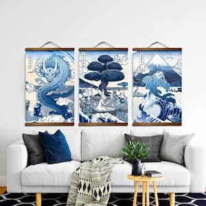Puede incluir: Conjunto de tres impresiones artísticas murales de estilo japonés en azul y blanco. Cada impresión presenta una escena diferente: un dragón, un árbol con figuras y una carpa koi con un fondo montañoso. Las impresiones están montadas en colgadores de madera.