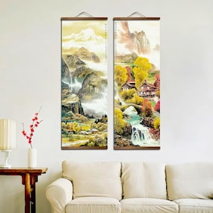 May include: Two vertical landscape paintings with wooden hanging rods. The artwork depicts a mountain scene with waterfalls, trees, and buildings. The color palette includes greens, yellows, and browns, with a soft, ethereal quality.