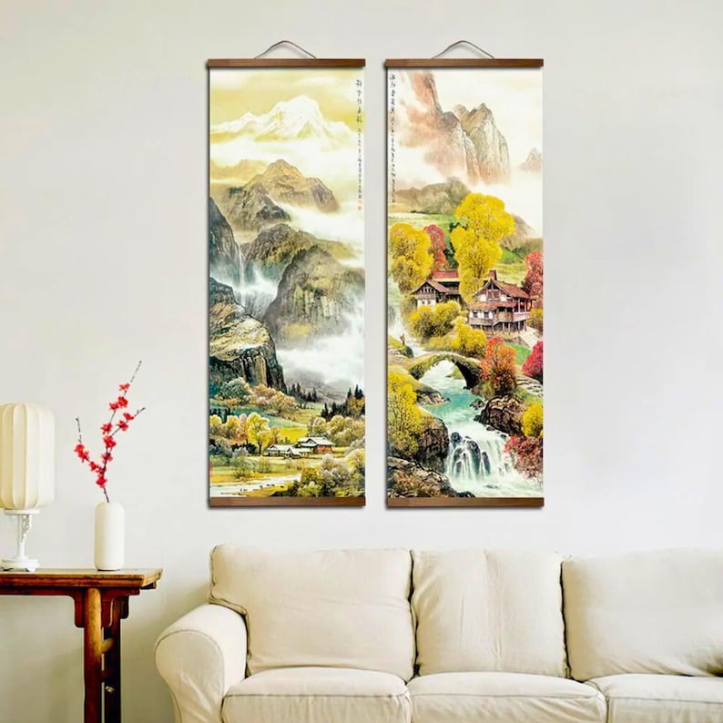 May include: Two vertical landscape paintings with wooden hanging rods. The artwork depicts a mountain scene with waterfalls, trees, and buildings. The color palette includes greens, yellows, and browns, with a soft, ethereal quality.