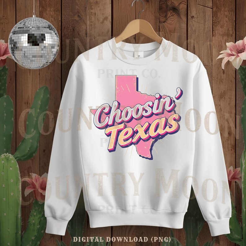 May include: White crew neck sweatshirt featuring a pink outline of Texas and the words "Choosin' Texas" in a retro font. The design has a vintage aesthetic. The sweatshirt is hanging on a wooden hanger.