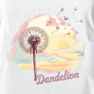 May include: White t-shirt featuring a detailed illustration of a dandelion with seeds blowing in the wind. The design includes a pastel sunset background and the word "Dandelion" in a script font.