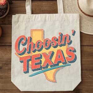 Op de afbeelding: Cr&egrave;mekleurige draagtas met de tekst "Choosin' TEXAS" in retro-lettertype, met een oranje en gele omtrek van Texas. De tas heeft twee handvatten en is gemaakt van canvas.