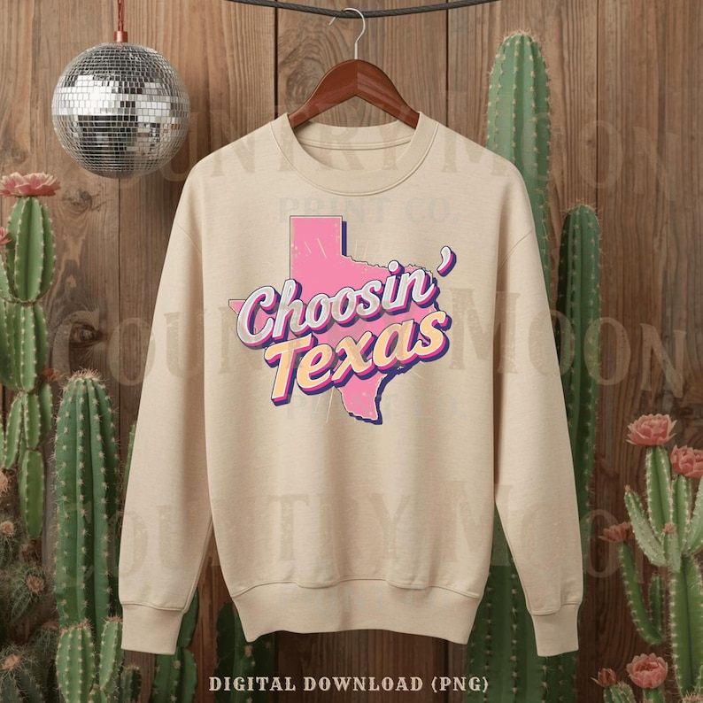 May include: A beige sweatshirt featuring the text "Choosin' Texas" in pink and yellow, with a pink outline of Texas. The sweatshirt is displayed on a wooden hanger, set against a backdrop of cacti and a disco ball.