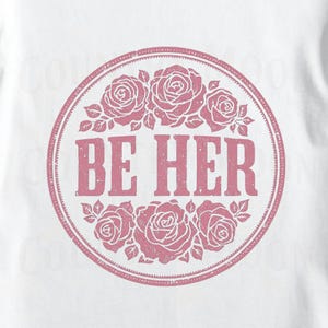 May include: White t-shirt featuring a circular design with the text "BE HER" in a distressed font. The design is surrounded by a ring of pink roses and leaves, creating a floral and feminine aesthetic.