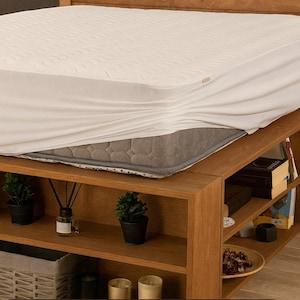 Può includere: Un coprimaterasso impermeabile bianco copre parzialmente un materasso grigio su un letto con struttura in legno. Il letto ha una testiera con un design geometrico e un'unità di stoccaggio integrata. Il testo sull'immagine recita "WATERPROOF MATTRESS PROTECTOR".