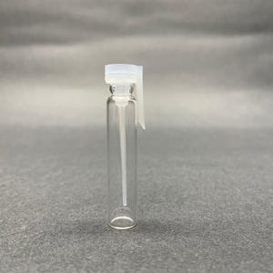 Puede incluir: Un vial de muestra de perfume de vidrio transparente con un pulverizador y una tapa de plástico blanco. El vial es cilíndrico y vacío, con un tubo largo y delgado que se extiende desde el pulverizador. El fondo es gris sólido.