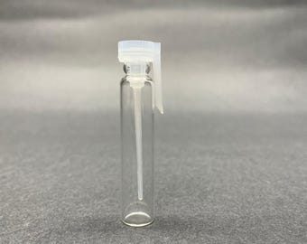 Op de afbeelding: Een heldere glazen parfummonsterfles met een witte plastic verstuiver en dop. De flacon is cilindrisch en leeg, met een lange, dunne buis die zich uitstrekt vanaf de verstuiver. De achtergrond is effen grijs.