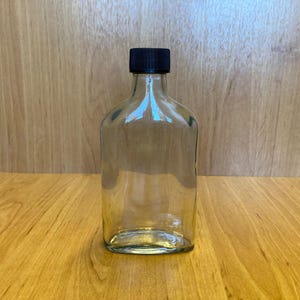 Peut inclure: Une flasque en verre transparent avec un bouchon noir. La flasque a une forme rectangulaire avec des coins arrondis. L'arrière-plan est une surface en bois.