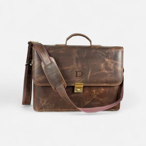May include: A brown leather briefcase with a top handle and a detachable shoulder strap. The bag features a flap closure with a gold-colored clasp and a debossed "D" logo. The leather has a distressed finish.