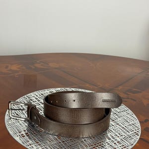 May include: A brown leather belt with a silver buckle and a textured surface. The belt is coiled on a white, patterned surface. The belt is a neutral color and could be worn with many outfits.
